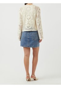 Koronkowy kardigan w kolorze kremowym z kwiatowymi wzorami, połączony z dżinsową spódnicą w kształcie litery A. Całość dopełniają sandały na obcasie. Zaprezentowane z tyłu.