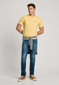 T-shirt di cotone gialla con scollo rotondo, abbinata a jeans blu. Una camicia a righe è legata in vita. Scarpe marroni e bianche completano il look.