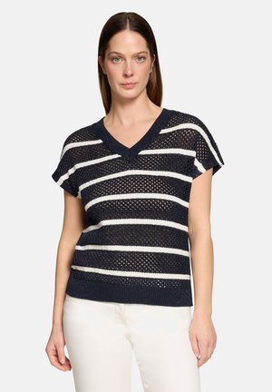 Femme portant un haut en maille à manches courtes rayé bleu marine et blanc avec un col en V et un pantalon blanc, debout devant un fond uni.