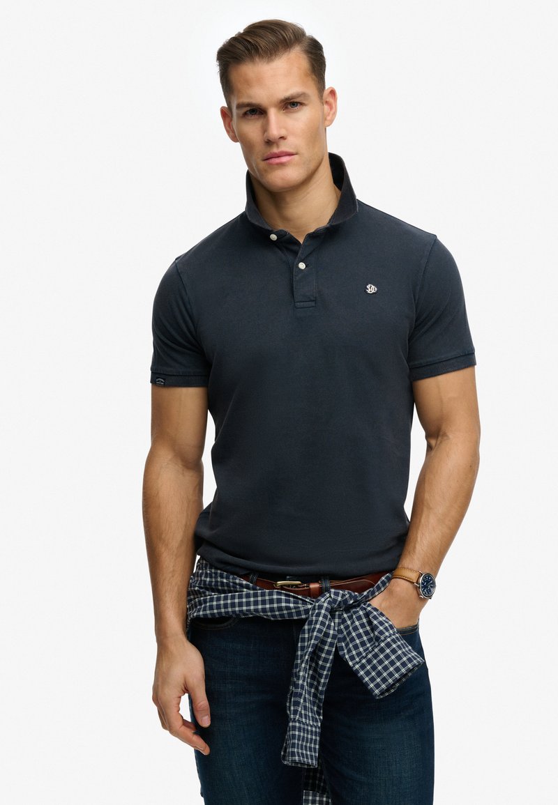 Polo azul marino con cuello, mangas cortas y un logotipo en el pecho; se lleva con vaqueros oscuros y una camisa azul a cuadros atada a la cintura.