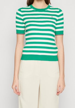 Vrouw draagt een groen-wit horizontaal gestreept korte mouw gebreide top met crèmekleurige broek met hoge taille, staand tegen een effen achtergrond.
