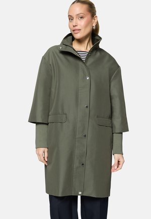 Femme portant un manteau mi-long vert olive avec des manches courtes larges et des boutons-pression, par-dessus une chemise à rayures à manches longues et un pantalon foncé.