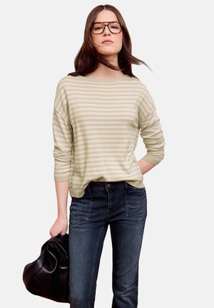 Femme aux longs cheveux bruns et lunettes, portant un pull beige à rayures et un jean foncé, tenant une veste en cuir noire dans une main.