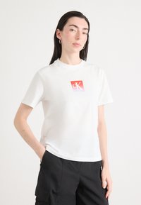 Witte katoen t-shirt met een vierkant rood logo en een gestileerde "ck." Korte mouwen, ronde halslijn, gedragen met zwarte cargobroeken.