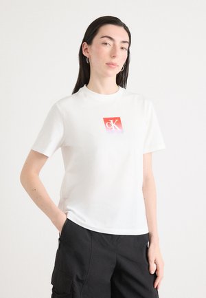 Hvit bomull t-skjorte med et kvadratisk rødt logo med en stilisert "ck." Korte ermer, rund hals, brukt med svarte cargo-bukser.