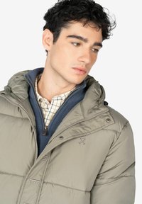 Giacca puffer in verde oliva, con cappuccio staccabile, chiusura con zip e cuciture a vista. Indossata sopra una giacca blu con zip e una camicia a quadri.