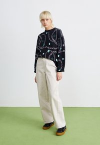 Pullover preto com um design de corrente e detalhes azuis, combinado com calças largas bege e sapatilhas pretas com sola em borracha sobre um pavimento verde.
