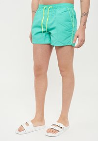 Shorts de bain turquoise avec cordon jaune, poches latérales et tissu léger ; associés à des sandales blanches à enfiler. Design minimaliste.