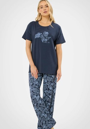 Mujer rubia de pie con pijama azul marino con estampado de conchas, camiseta de manga corta y pantalones anchos a juego sobre un fondo liso.