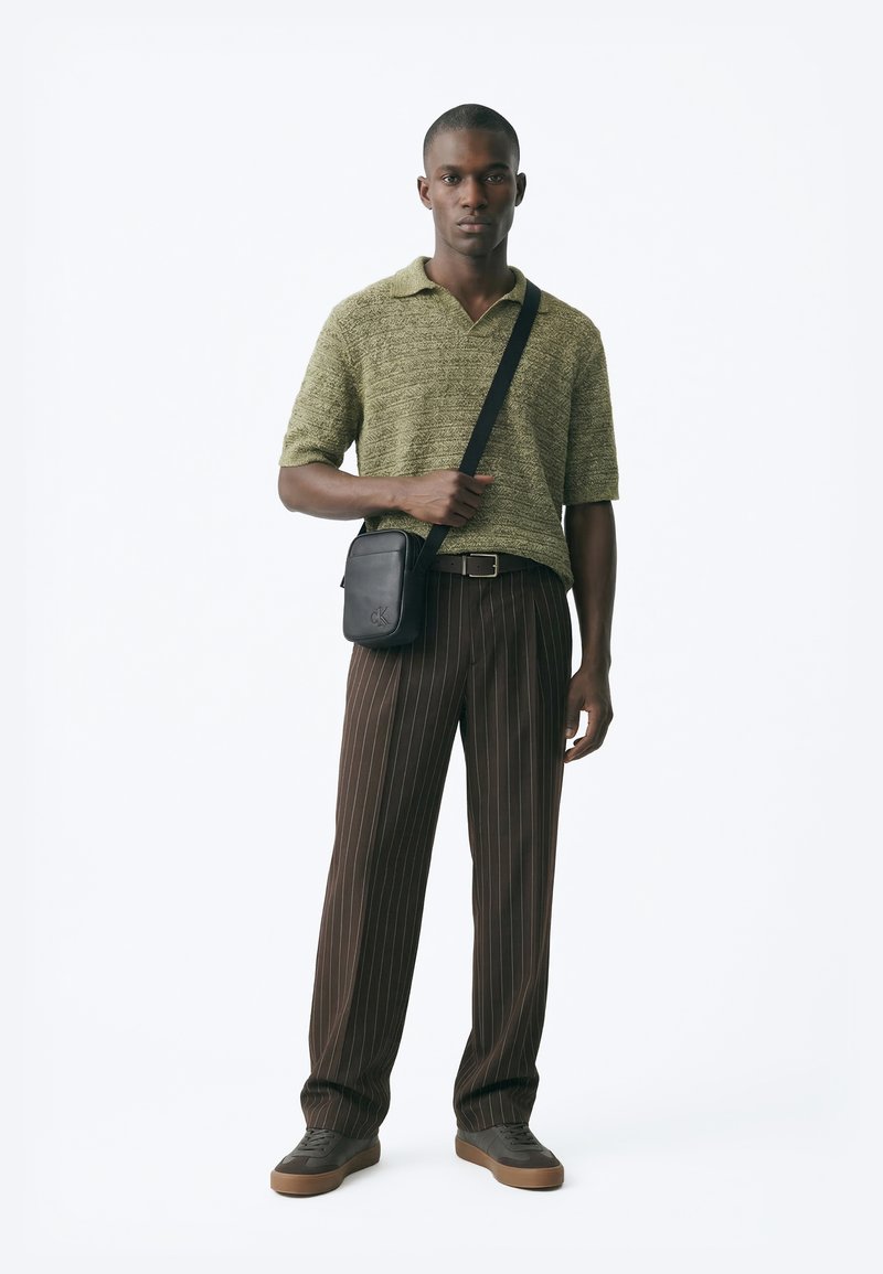 Joven con polo de manga corta texturizado verde, pantalones marrón con rayas finas, bolso bandolera negro y zapatillas marrones, posando frente a fondo blanco.