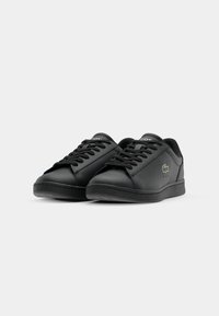 Sorte syntetiske sneakers med slankt design, runde tæer, snørebånd foran, subtile blå accenter og en tekstureret gummisål. Lacoste-logo på siden.
