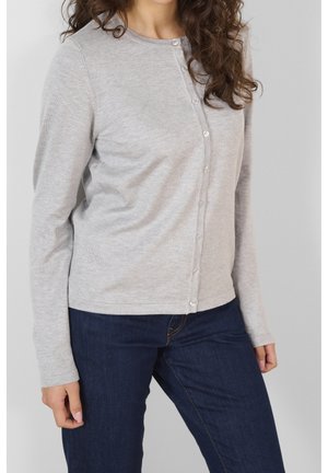 Femme portant un cardigan boutonné gris clair et un jean bleu foncé, debout devant un fond uni gris clair.