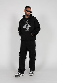 Schwarzer Hoodie mit Frontgrafik, kombiniert mit schwarzen Cargo-Hosen mit seitlichen Taschen und schwarz-weißen Sneakers. Strukturierter Stoff, lockere Passform.