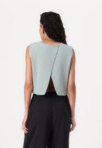 Donna con lunghi capelli ricci che indossa un top azzurro chiaro corto senza maniche con spacco sulla schiena e pantaloni neri a vita alta e gamba larga, in piedi davanti a uno sfondo bianco.