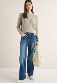 Femme portant un pull en maille beige, un jean large bleu, des baskets blanches, tenant un grand sac fourre-tout crème, debout devant un mur clair uni.