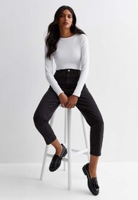 Maglia bianca a coste a maniche lunghe abbinata a jeans neri a vita alta e mocassini neri. Il modello è seduto su uno sgabello bianco contro uno sfondo chiaro.