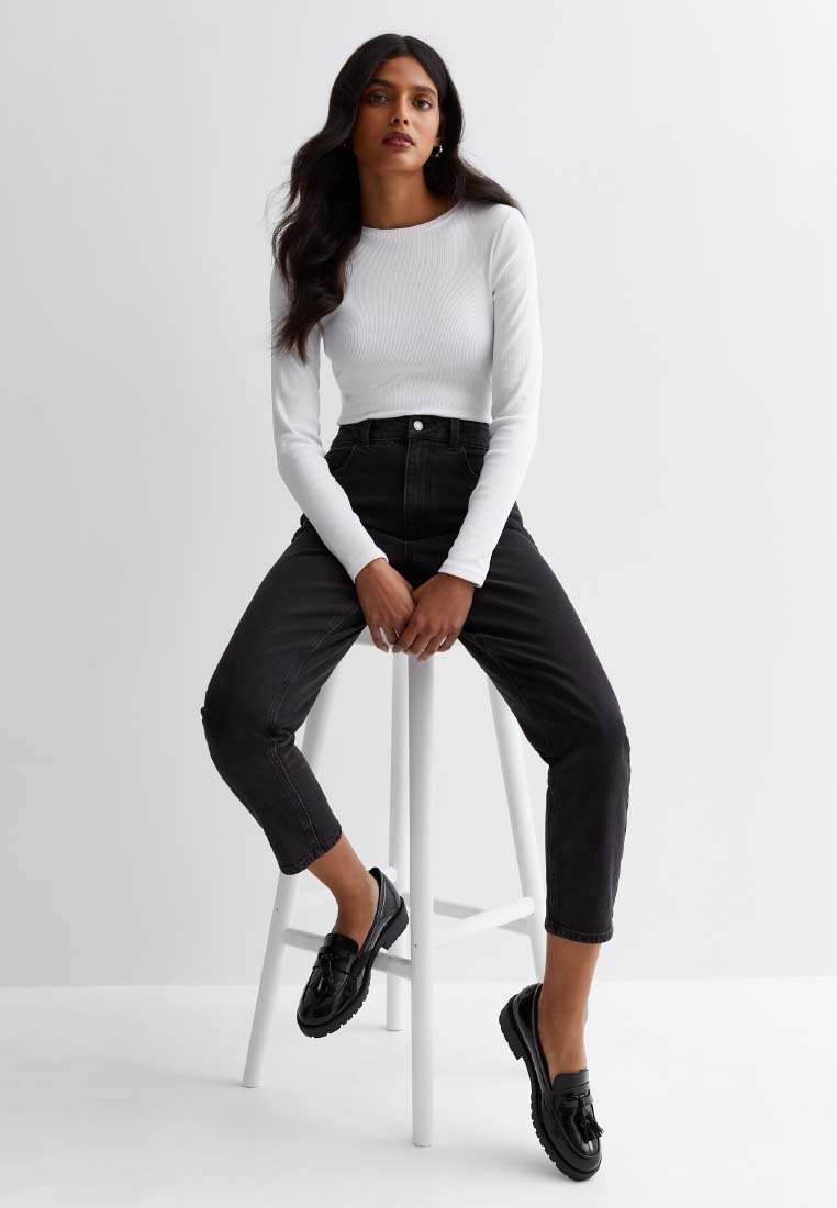 Maglia bianca a coste a maniche lunghe abbinata a jeans neri a vita alta e mocassini neri. Il modello è seduto su uno sgabello bianco contro uno sfondo chiaro.