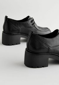 Schwarze Lederschuhe mit runder Zehenform, flachen Schnürsenkeln und einem dicken, blockartigen Absatz mit strukturiertem Unterboden. Glatte Oberfläche mit dezenten Nähten.