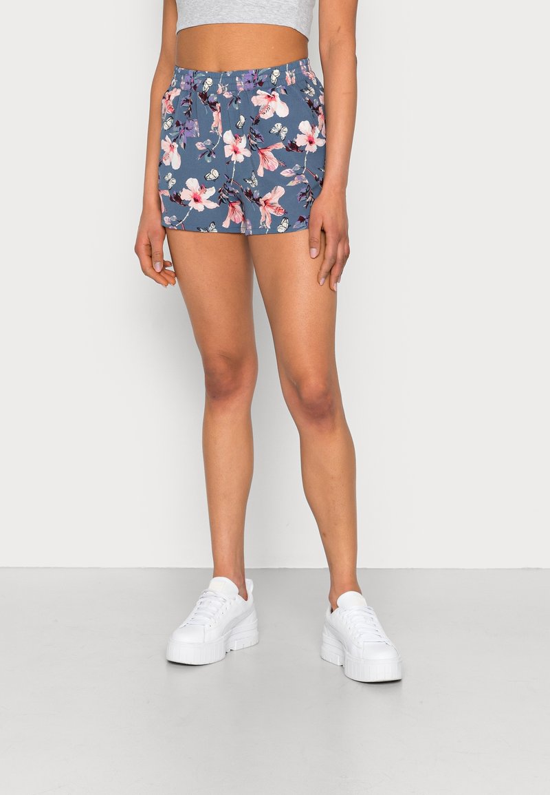 Femme portant un short bleu à imprimé floral et des baskets à plateforme blanches, debout devant un fond blanc uni.