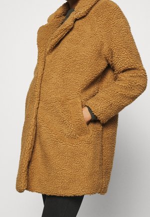 Person iført en mellemlang, lysebrun, tekstureret teddyfrakke med sidelommer, den ene hånd i lommen, over sorte bukser.