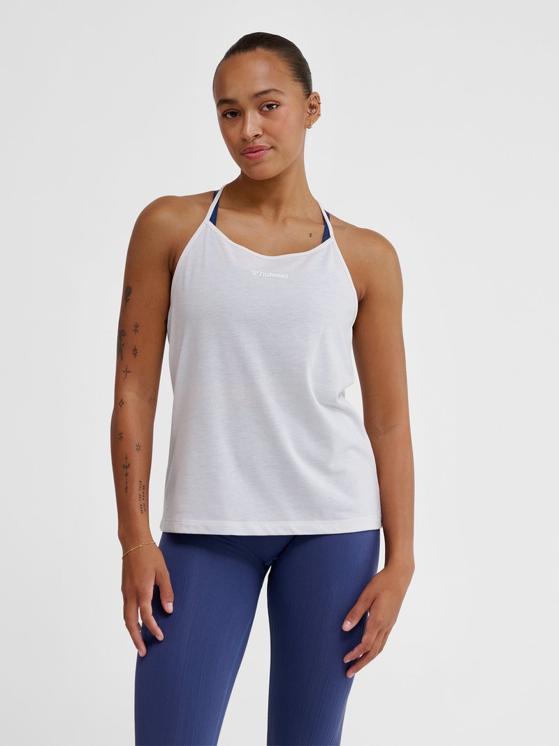 Vrouw met naar achteren gebonden haar, gekleed in een wit mouwloos sporttopje en blauwe leggings, staand tegen een effen lichte achtergrond.