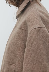 Brauner Wollmischmantel mit strukturiertem Kragen, strukturiertem Stoff und einer Seitentasche. Der Stoff hat eine weiche, gebürstete Oberfläche.