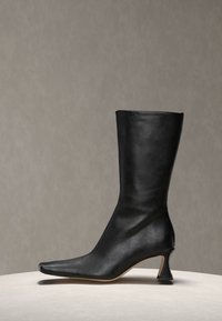 Μαύρη δερμάτινη γυναικεία μπότα μέσης γάμπας με μυτερό δάχτυλο και κυρτό τακούνι block, τοποθετημένη σε στρογγυλή ελαφριά βάση μπροστά σε γκρι φόντο.