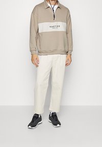 Beige tröja med halv dragkedja och ett vitt horisontellt rand med texten "KAOTIKO". Matchad med ljusgräddafärgade byxor och svarta sneakers.