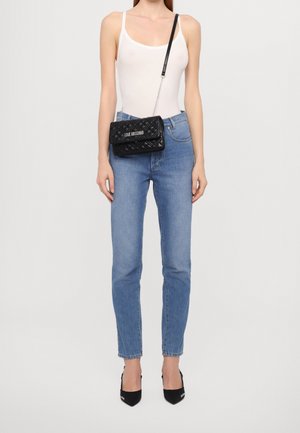 Vrouw die een wit tanktop draagt, lichtblauwe hoge taille jeans, zwarte gewatteerde schoudertas met het logo "Love Moschino" en zwarte puntige hakken.