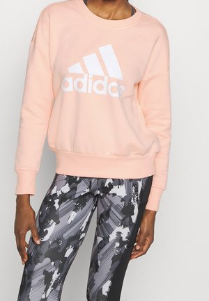 Person trägt ein hellrosa Adidas-Sweatshirt kombiniert mit schwarzen, weißen und grauen Leggings mit abstraktem Muster, steht vor einem einfarbigen Hintergrund.