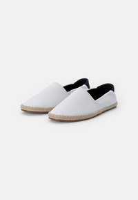 Hvite kanvas slip-on sko med spiss tå, jute-detaljering rundt sålen og en kontrasterende mørk ankelkant.
