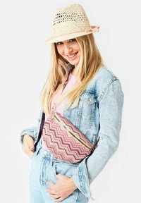 Gewebte Stoff-Crossbody-Tasche mit einem Chevron-Muster in Pink und Lila, ausgestattet mit einem goldenen Reißverschluss. Das Modell trägt eine Jeansjacke und einen Strohhut.
