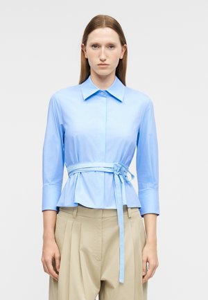 Kobieta ubrana w jasnoniebieską bluzkę z długim rękawem i paskiem wiązanym w talii oraz beżowe plisowane spodnie, stoi na jednolitym tle.