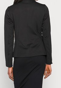 Veste noire cintrée avec manches longues, dos structuré et sans ornements, associée à une jupe noire côtelée. Tissu lisse et ajusté.