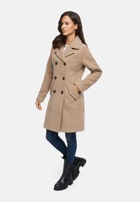 Cappotto beige doppiopetto con bottoni neri e tasche laterali. Il cappotto ha un colletto a revers ed è abbinato a jeans scuri e stivaletti neri.