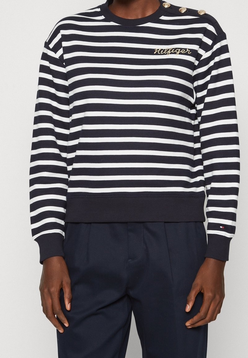 Personne portant une chemise à manches longues rayée bleu marine et blanche avec des boutons dorés sur l'épaule et "Hilfiger" brodé sur la poitrine.