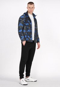 Camo-jakke i blå og sort stof med lynlås foran, hætte og elastiske manchetter, parret med sorte bukser og hvide sneakers.
