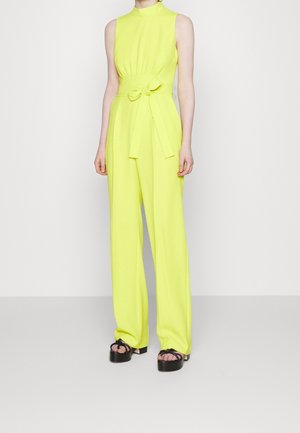 Ærmeløs neon gul jumpsuit med høj hals og bånd i taljen, båret med sorte plateausandaler, på en person, der står mod en hvid baggrund.