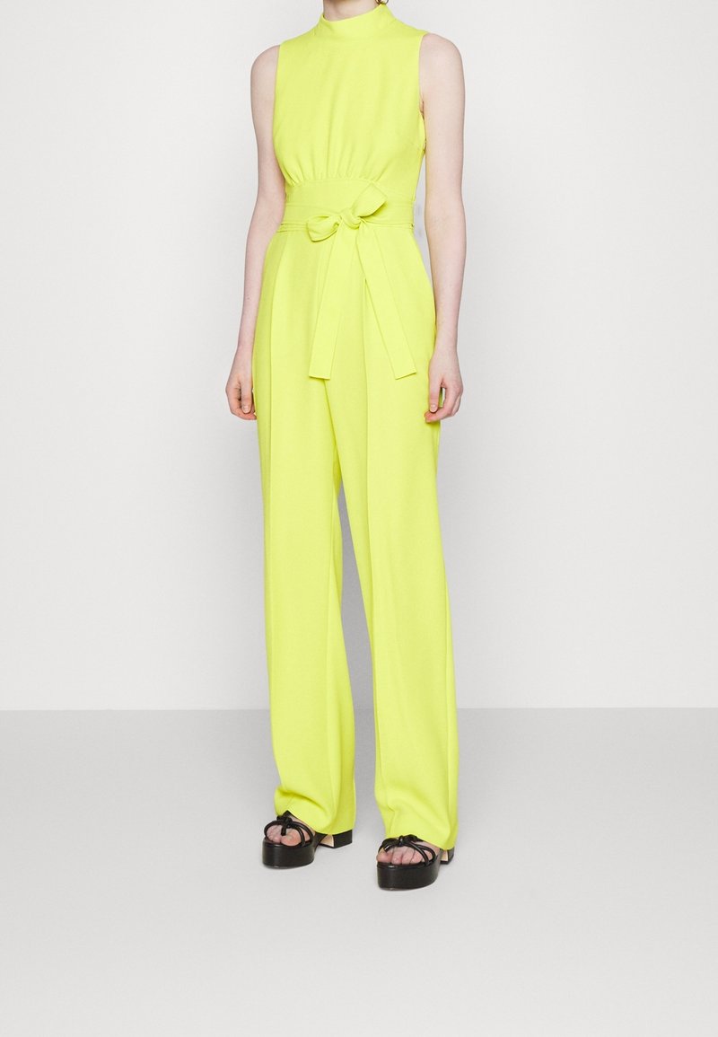 Combinaison sans manches jaune fluo avec col montant et ceinture à la taille, portée avec des sandales plateformes noires, sur une personne debout sur fond blanc.
