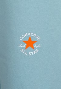 Ljusblått tyg med en vit cirkulär logotyp med en orange stjärna och texten "CONVERSE Chuck Taylor ALL STAR" i en sportig design.