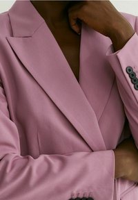 Personne portant un blazer rose à boutons noirs, bras gauche croisé, main droite touchant le menton sur un fond neutre.