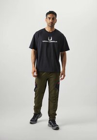Černé bavlněné tričko s bílým logem "Under Armour", které je kombinováno s olivově zelenými kalhotami s červenými a navy akcenty a černými detaily.