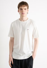 T-shirt blanc en coton avec un col rond, des manches courtes et un petit logo sur la poitrine gauche. Texture lisse et coupe décontractée.