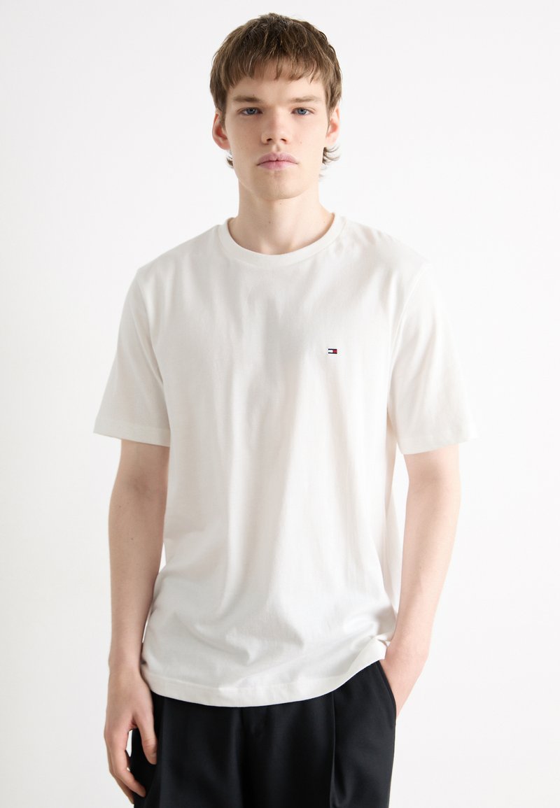 T-shirt blanc en coton avec un col rond, des manches courtes et un petit logo sur la poitrine gauche. Texture lisse et coupe décontractée.