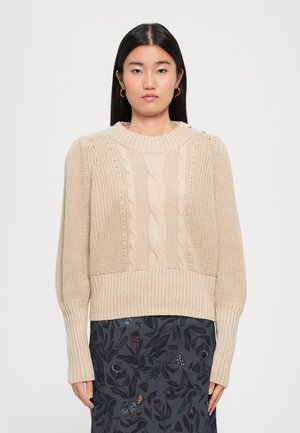 Woman wearing a beige cable-knit sweater with long ribbed sleeves and a dark skirt with a floral pattern, standing against a plain background.
