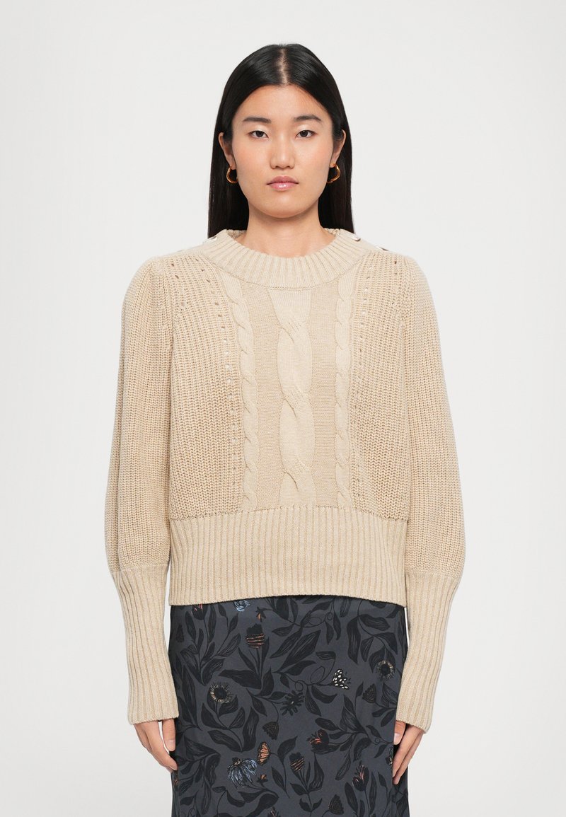 Woman wearing a beige cable-knit sweater with long ribbed sleeves and a dark skirt with a floral pattern, standing against a plain background.