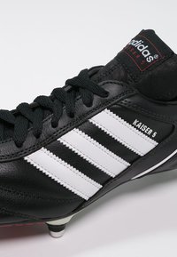 Scarpa da calcio in pelle nera con tre strisce bianche, lacci neri e linguetta con bordatura rossa etichettata "adidas Kaiser 5". Suola con tacchetti.