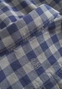 Tessuto a quadri blu e grigio con una consistenza morbida. Presenta un logo ricamato e bordi cuciti, evidenziando il design e il contrasto dei colori.