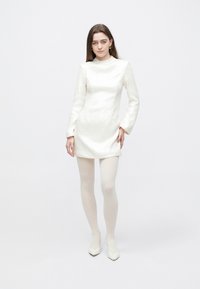 Wit mini-jurkje met lange mouwen, gestructureerde stof, hoge hals en een aansluitend silhouet; gecombineerd met witte panty's en puntige hakken.