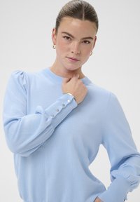 Pull en maille bleu clair avec manches bouffantes et détail de boutons aux poignets. Texture douce et coupe ajustée. Fond uni.